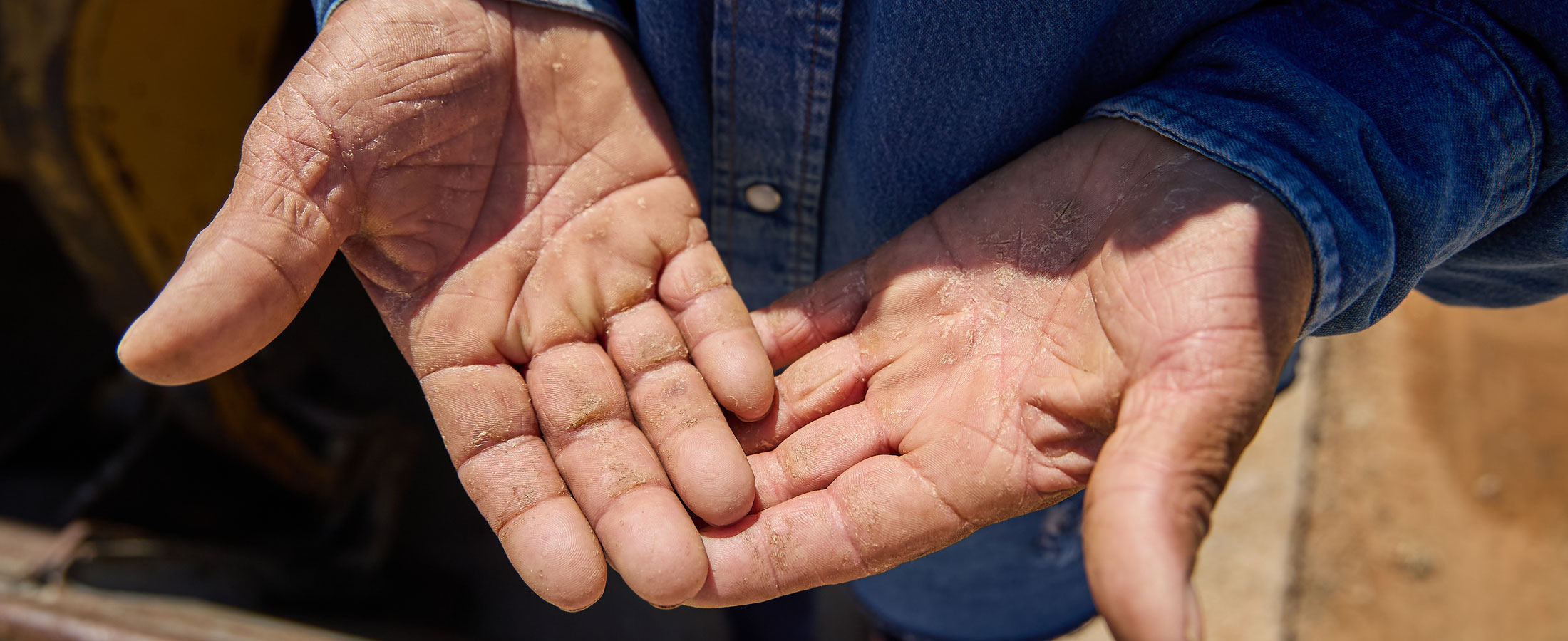 Close-up of a person’s open hands showing worn, calloused palms, wearing a denim work shirt, standing outdoors at a construction site.